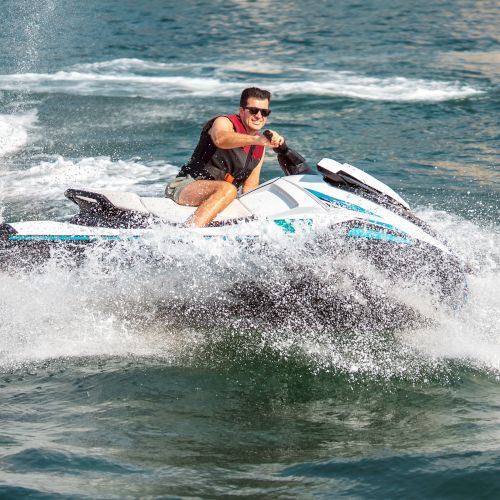 A man rides a jet ski through churning waves, smiling in the spray as he speeds across open water.