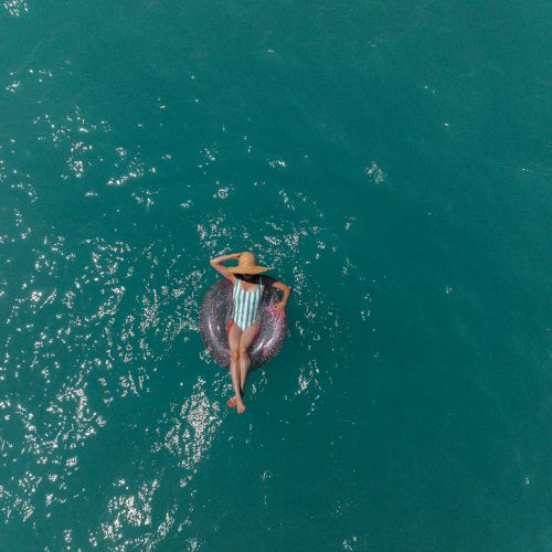 A person in a striped swimsuit floats on their back in a sparkling turquoise ocean, wearing a straw hat.