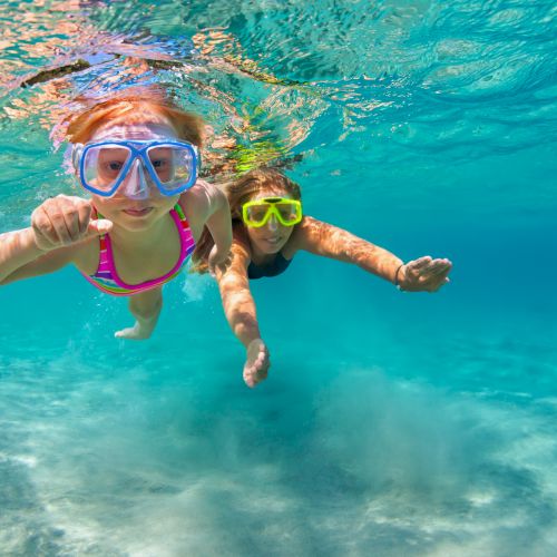 Two kids snorkel in clear turquoise water, wearing colorful masks and waving as they swim together.