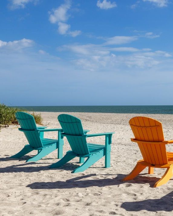 Colorful Adirondack chairs sit on a sandy beach overlooking the calm ocean under a bright blue sky.