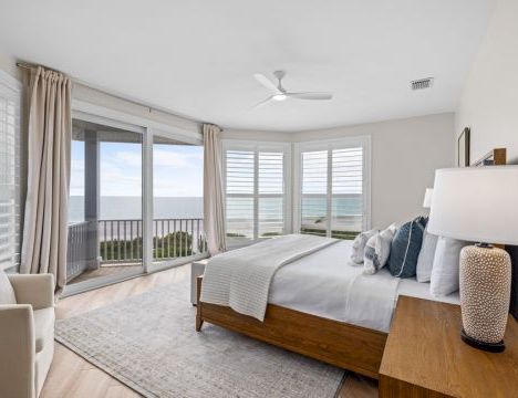 A bright modern bedroom with a large bed, ceiling fan, and floor-to-ceiling windows showing an ocean view.