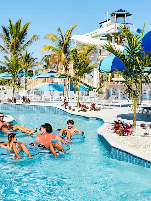 Sunny resort pool with palm trees, blue slides, and kids and adults floating in inner tubes, soaking up vacation vibes.