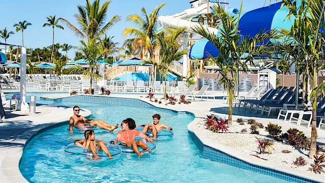 Sunny resort pool with palm trees, blue slides, and kids and adults floating in inner tubes, soaking up vacation vibes.