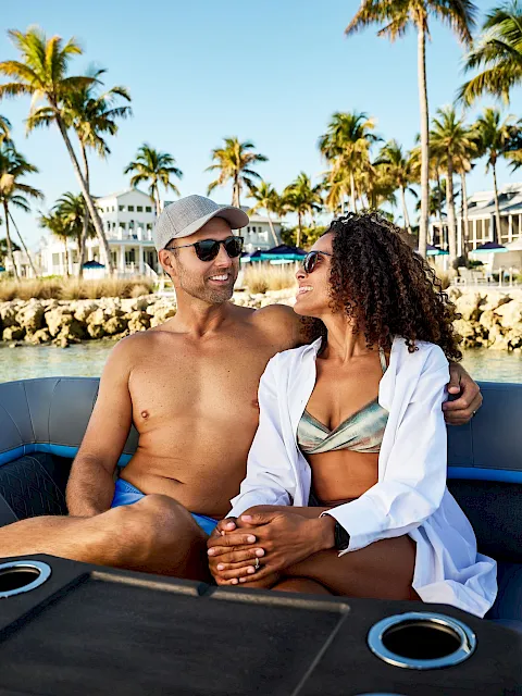 A sunny beach getaway on a boat: a smiling couple snuggles together with palm trees and a resort marina in the background.