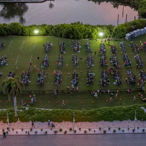 An aerial view of a large outdoor game or training event with many people in organized groups on a grassy field by a river.