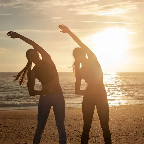 Two people dance on a beach at sunset, arms raised above the waves behind them.