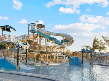 A sunny water park with a large multi-lane blue waterslide, a splash pool, and palm trees under a bright blue sky.