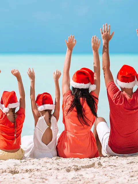 Four people in red outfits and Santa hats sit on the sand with their arms raised against a blue sea background.